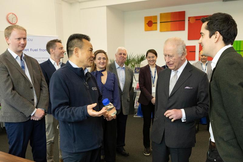 King Charles III stands with researchers at Oxford University Science Park, listening as a scientist explains a sample bottle, with other team members gathered around.
