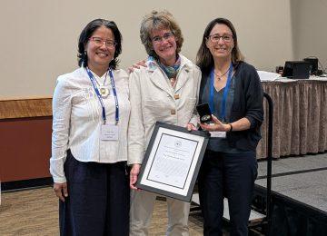 Prof Christiane Timmel receives the International EPR Society Medal and a certificate, standing between Profs Songi Han and Marina Bennati. They are all smiling. 