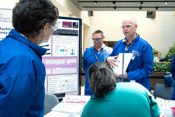 A woman in a blue lab coat and safety specs speaks to two men wearing the same.