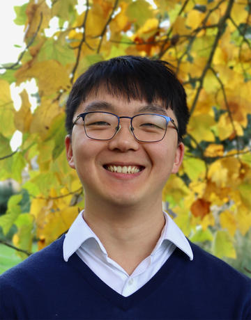 Headshot of Shurui Miao, a man with dark hair and glasses, smiling and wearing a navy sweater and white collared shirt. The background is yellow autumn leaves in a park.