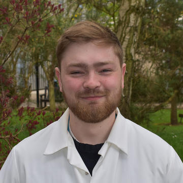 Head and shoulders photo of Theo Tanner, a man with short hair and a beard wearing a labcoat and smiling, stood in front of trees.