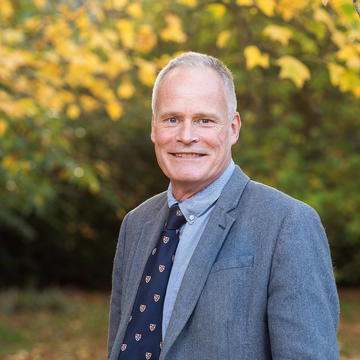 Mcgradyj Portrait photo of a man with short grey hair wearing a suit and smiling. The background is autumn leaves.