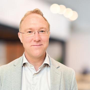 A head and shoulders portrait of Prof Andrew Baldwin, a man with light coloured hair and glasses wearing a grey suit, with an office background.