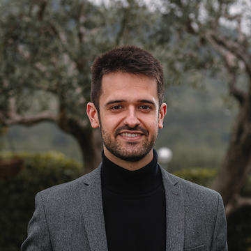 A head and shoulders photo of Pol Hernandez Llado, a man with dark hair and a beard smiling, standing in front of trees, wearing a black turtleneck and a grey jacket.