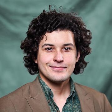A man with curly brown mid-length hair wearing a green shirt and a brown jacket stands in front of a green neutrla backdrop.