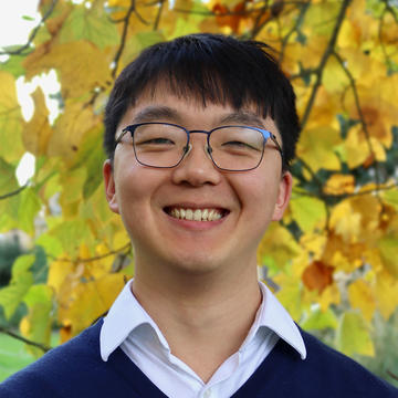 Headshot of Shurui Miao, a man with dark hair and glasses, smiling and wearing a navy sweater and white collared shirt. The background is yellow autumn leaves in a park.
